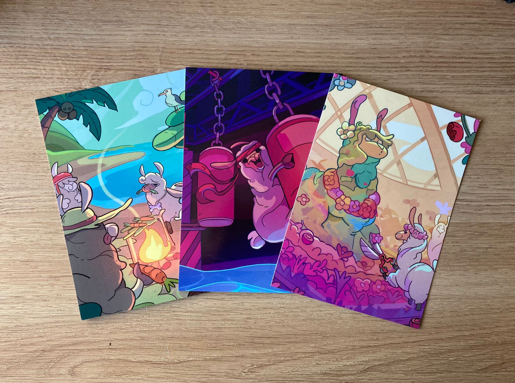 A photograph of three postcards featuring colourful illustrations of llamas in reality TV situations. One is of a group of llamas roasting carrots over a fire on a tropical island, another shows a gleeful llama swinging on a punch bag over a pool of water, and the last shows a llama in a flower crown trimming a huge llama topiary 